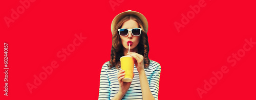 Portrait of stylish young woman drinking fresh juice wearing summer straw hat, sunglasses on red background