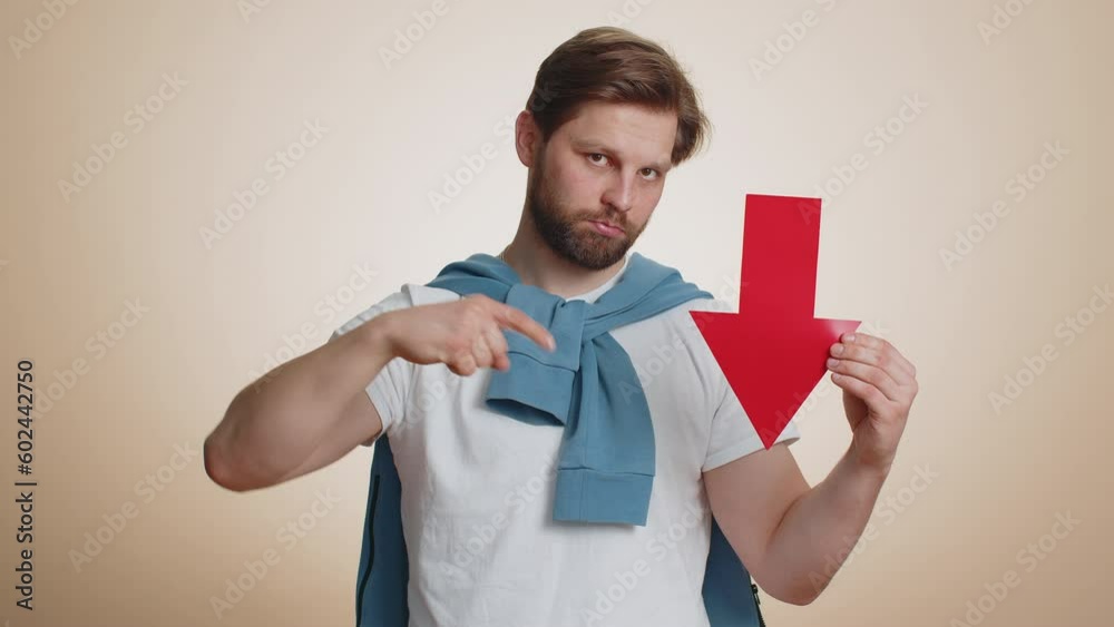 Upset caucasian man showing red arrow pointing down, concept of downgrade, unsuccessful business fall of stock market money exchange rate bankruptcy fail. Young guy isolated on beige studio background Upset caucasian man showing red arrow pointing down, concept of downgrade, unsuccessful business fall of stock market money exchange rate bankruptcy fail. Young guy isolated on beige studio background