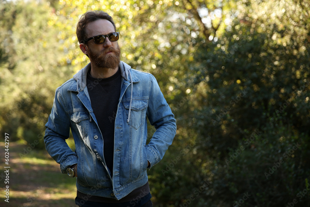 Trekking lover, adventurer, bearded Caucasian man with sunglasses posing in front of a blurry green background with soft natural light