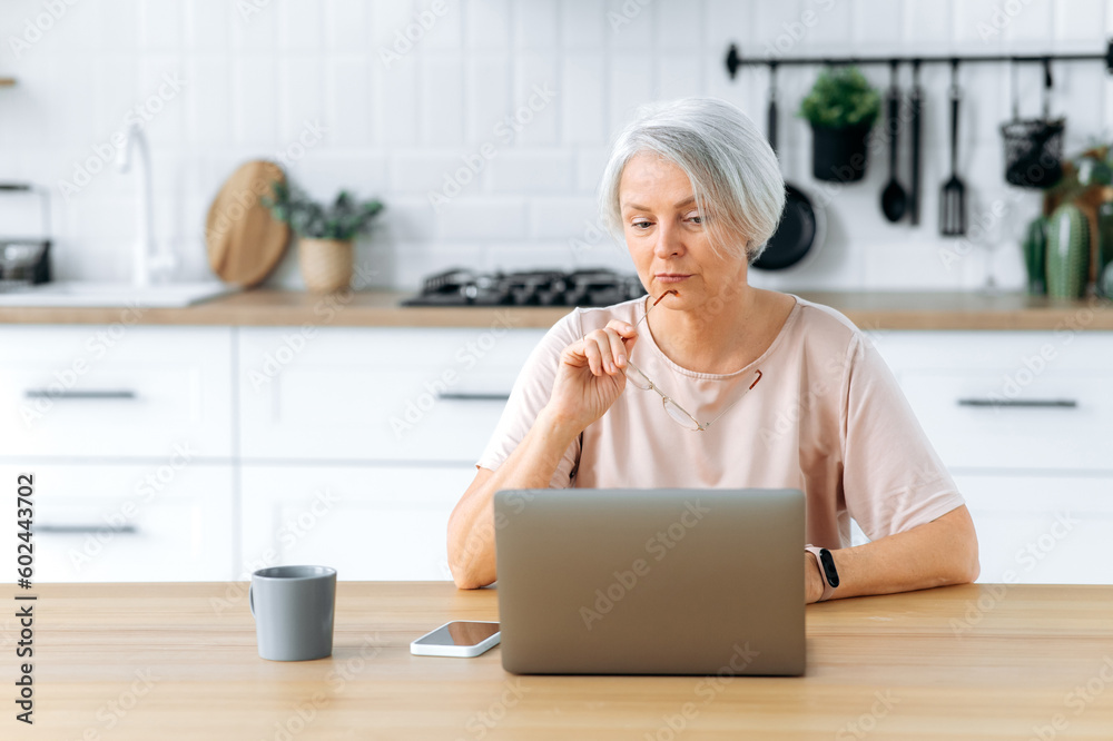 Puzzled caucasian gray haired lady uses a laptop to work online or search for information on the Internet, sits in the kitchen at a desk, looks confused at the screen, thinks about information, news