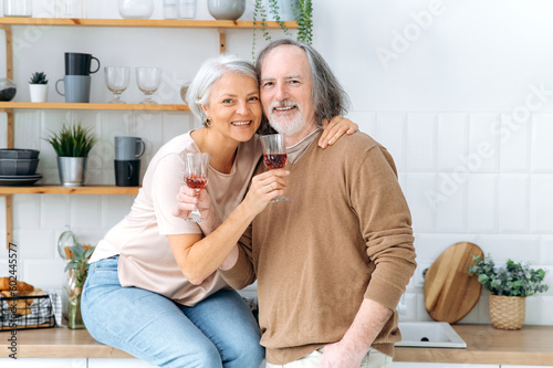 Anniversary. Happy pensioners with wine. Middle aged caucasian spouses, sit at home in the kitchen, hold glasses of red wine in their hands, celebrate an important date, anniversary, smile at camera