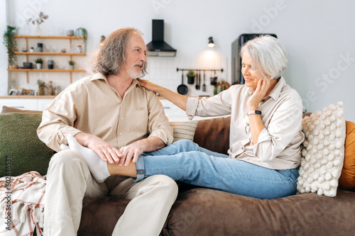 Caucasian gray haired pensioners, mature husband and wife, spend time together at home on the sofa in the living room, man massaging his beloved wife feet, they resting and relaxing together