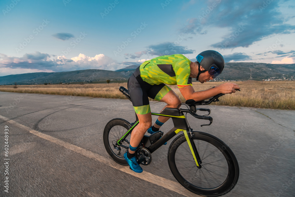 Fototapeta premium Triathlete riding his bicycle during sunset, preparing for a marathon. The warm colors of the sky provide a beautiful backdrop for his determined and focused effort.