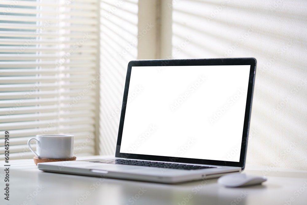 Mock up laptop computer with blank screen, mouse and cup of coffee on white working desk in bright office.