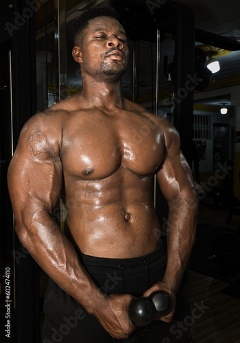 A black athlete is intensely exercising in the gym, strengthening their muscular build with a bare chested shoulder workout and sweating to achieve a healthy lifestyle.
