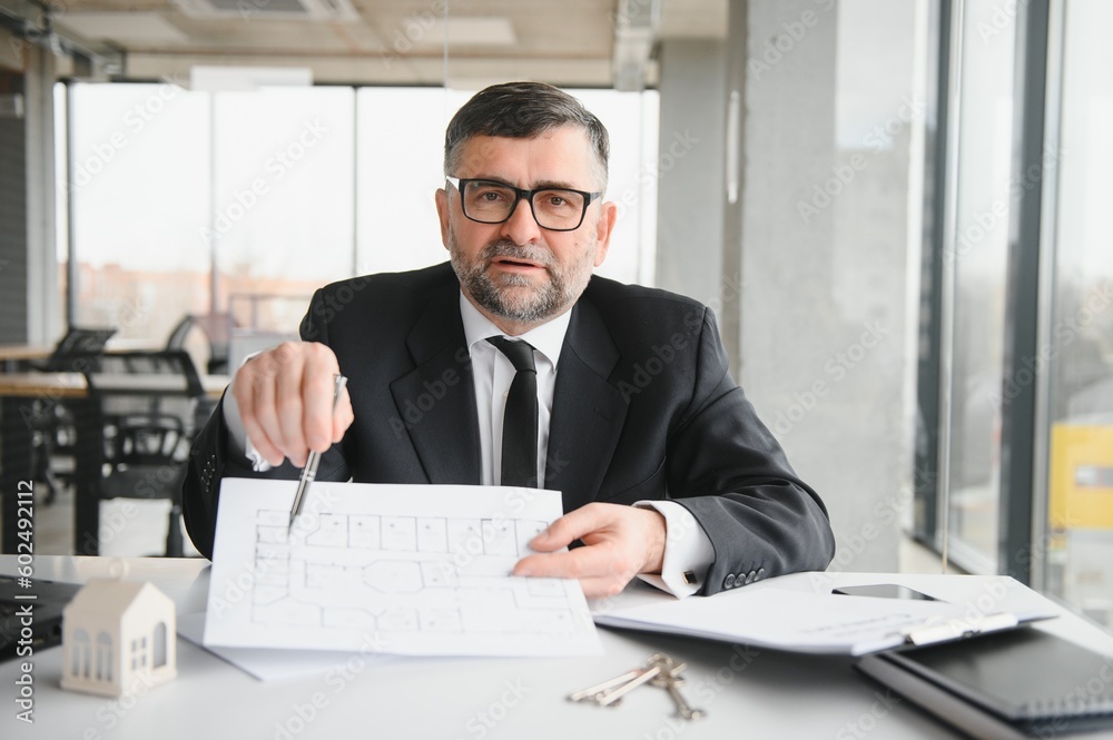 Realtor in suit sitting at desk in office. Realtor is showing apartment. Realtor in suit sitting at desk in office. Realtor is showing apartment.