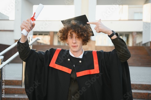 Low angle portrait of happy triumphant male graduate standing near university holding up diploma. From below of young handsome man proud of academic achievements celebrating college graduation