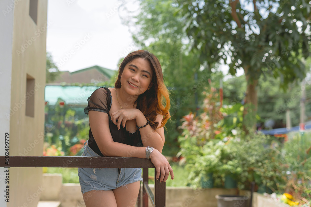Naklejka premium A pretty lady wearing a black blouse and jeans shorts is leaning on a rail, posing, hands on chest, smiling and looking sweetly at the camera. Plants and trees in the background.