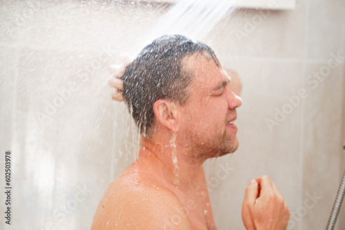 an ordinary man washes his hair in a shower in a bathroom with a shower head on the wall. Economical and simple interior for a house or hotel.