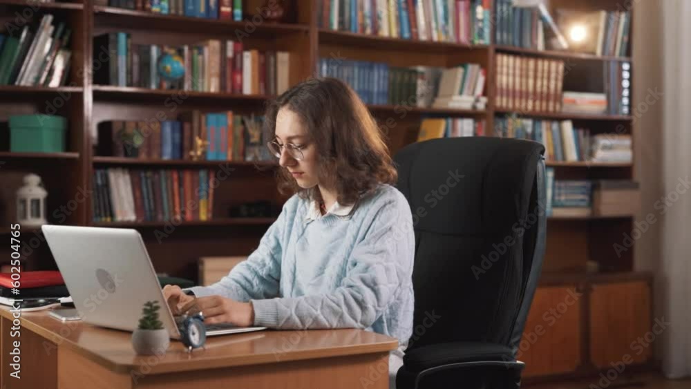 The girl is sitting at a table, typing something on her laptop. She might be working or replying to someone's message. In the end, she smiles magically, and there are lots of books around her. The girl is sitting at a table, typing something on her laptop. She might be working or replying to someone's message. In the end, she smiles magically, and there are lots of books around her.