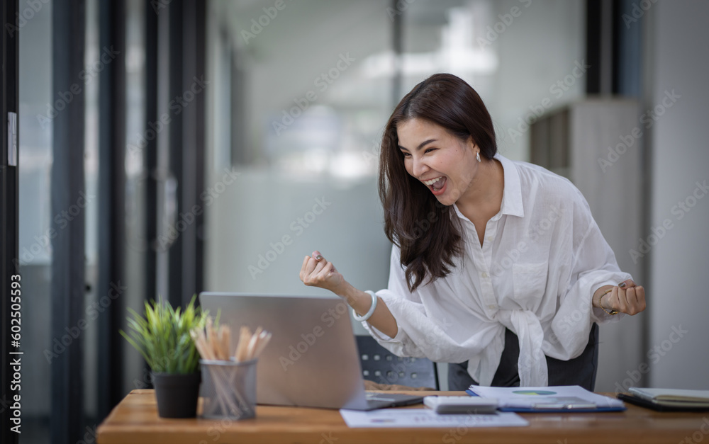 Excited Happy Asian young woman using phone and laptop sitting on a desk office in the day at office Excited Happy Asian young woman using phone and laptop sitting on a desk office in the day at office