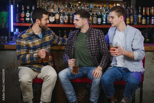 Three young men in casual clothes are smiling, holding bottles of beer while standing near bar counter in pub