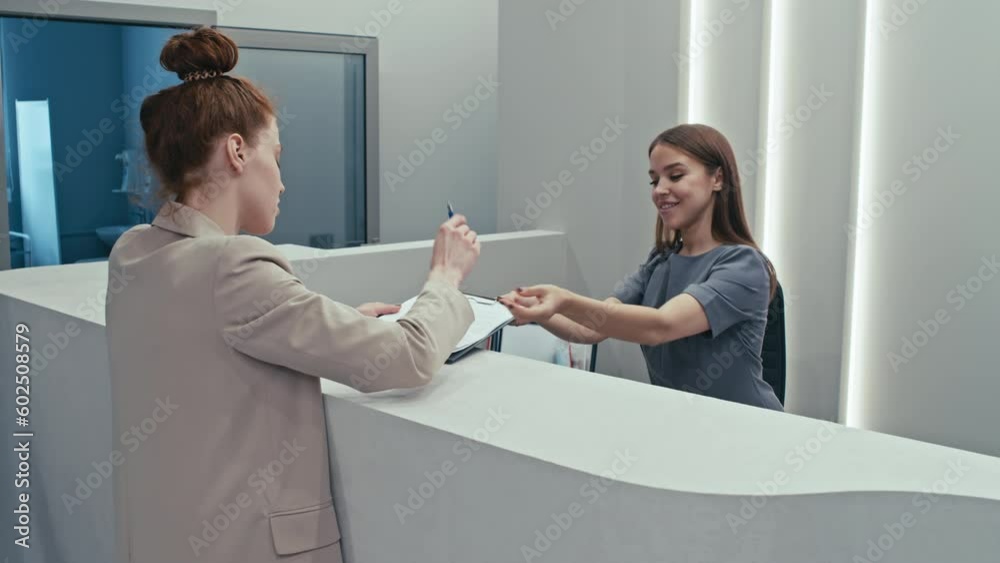 Young female patient having appointment in modern hospital signing agreement at reception desk