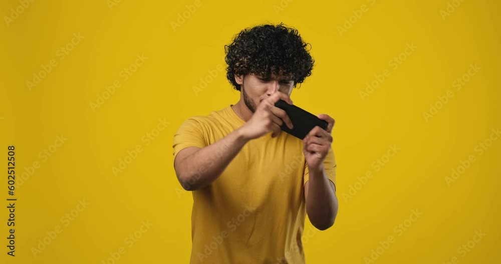 Smiling Happy Indian Man Gamer Using Mobile Phone Playing Game While Standing over orange studio background Smiling Happy Indian Man Gamer Using Mobile Phone Playing Game While Standing over orange studio background