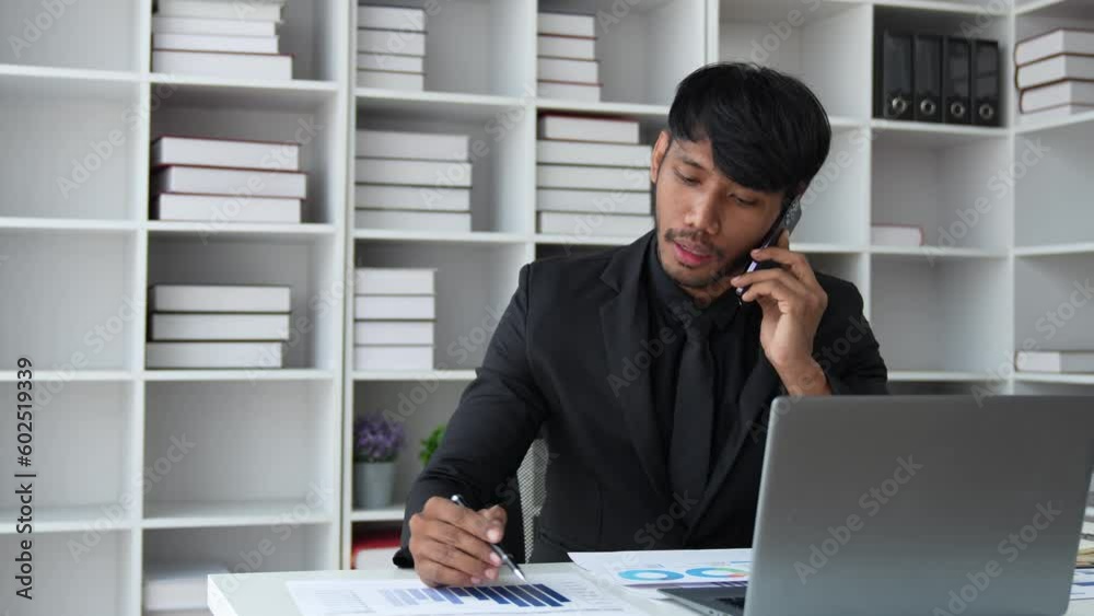 Handsome businessmen sitting at office desk in front of laptop hold mobile phone make pleasant business or informal call. Successful businessman looking at financial statistic shown on documents. Handsome businessmen sitting at office desk in front of laptop hold mobile phone make pleasant business or informal call. Successful businessman looking at financial statistic shown on documents.