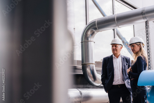 Businesswoman and senior businessman standing under metal pipe in factory photo