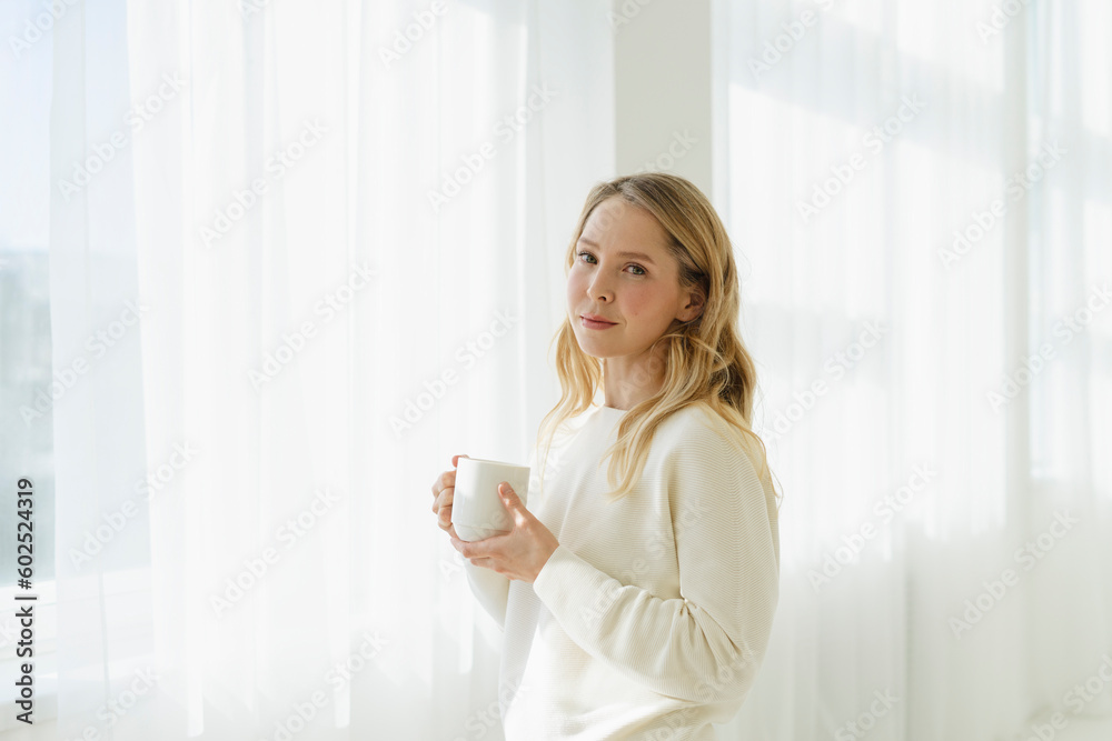 Blond woman holding coffee cup by translucent curtain Blond woman holding coffee cup by translucent curtain