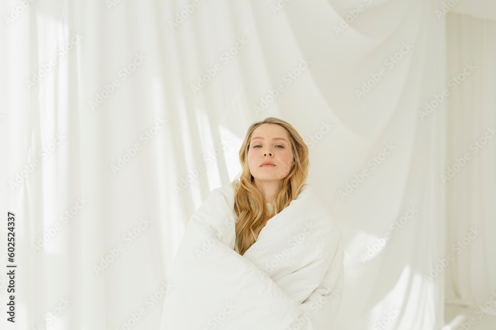Blond woman covered with white blanket in front of translucent curtain Blond woman covered with white blanket in front of translucent curtain