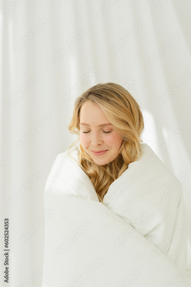Smiling woman with eyes closed wrapped in white blanket standing by translucent curtain Smiling woman with eyes closed wrapped in white blanket standing by translucent curtain