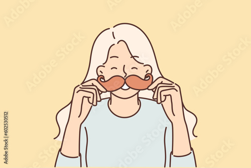 Cheerful girl puts cardboard mustache to face and laughs choosing funny mask for photo
