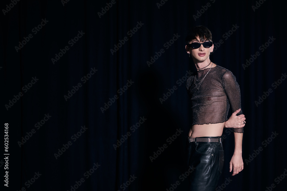 Trendy young gay man in sunglasses, sparking top and leather pants posing while celebrating lgbt community and pride month isolated on black Trendy young gay man in sunglasses, sparking top and leather pants posing while celebrating lgbt community and pride month isolated on black