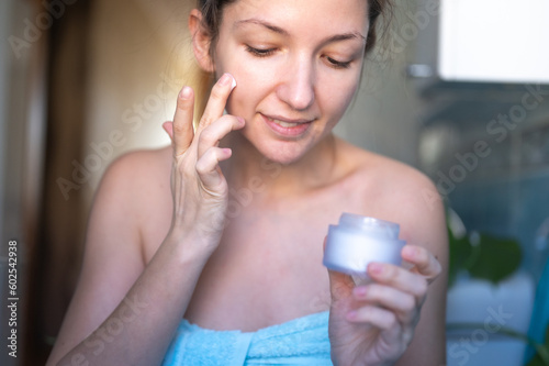 Young beatiful woman applies cream to her face. Skin care at home in the bathroom.