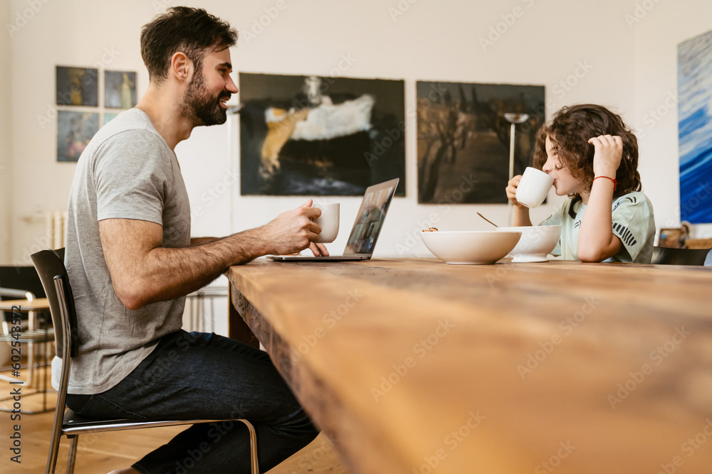 Man working on laptop while having breakfast with his son sitting at table