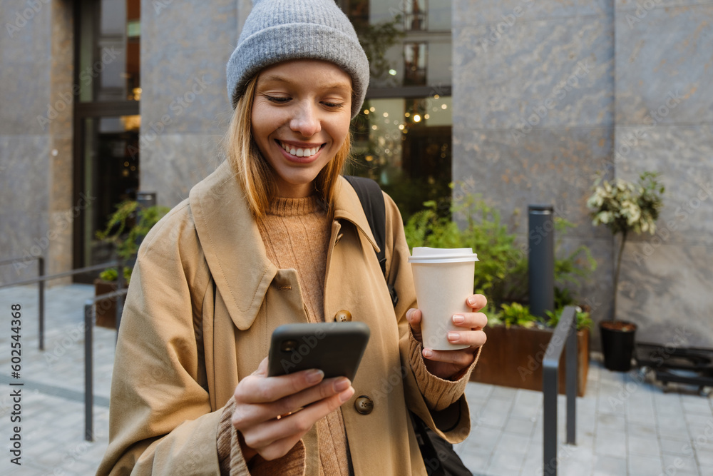Beautiful woman drinking coffee and using smartphone while walking outdoors through city street