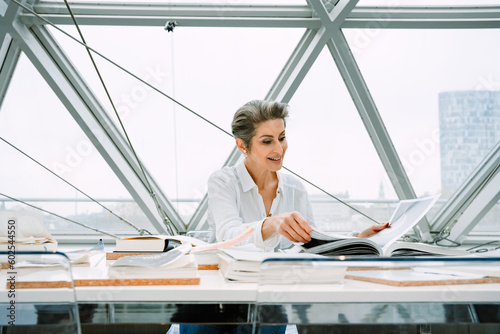 Smiling mature woman manager sitting at table with books while working in art gallery