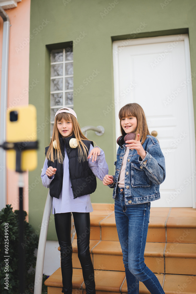 Young blonde sisters dance and record videos for social networks with their smartphones. The girls are standing in front of their house. Young blonde sisters dance and record videos for social networks with their smartphones. The girls are standing in front of their house.