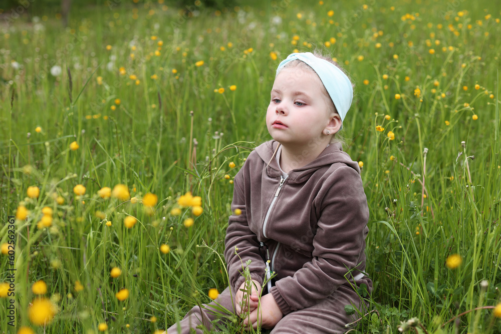 A little girl, 4 years old, sits in the big grass. A child on an uncut lawn. A child is playing on the street. Tall green grass in summer. Childhood in nature. Interested girl A little girl, 4 years old, sits in the big grass. A child on an uncut lawn. A child is playing on the street. Tall green grass in summer. Childhood in nature. Interested girl