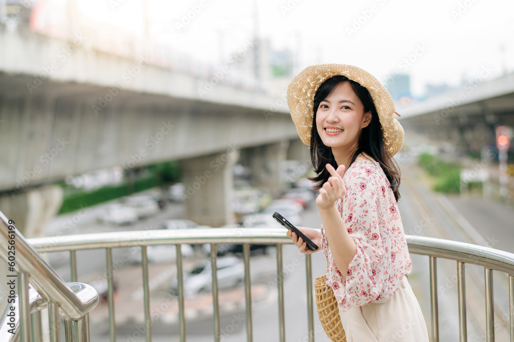 young asian woman traveler with weaving basket using mobile phone and standing on overpass with railway background. Journey trip lifestyle, world travel explorer or Asia summer tourism concept.