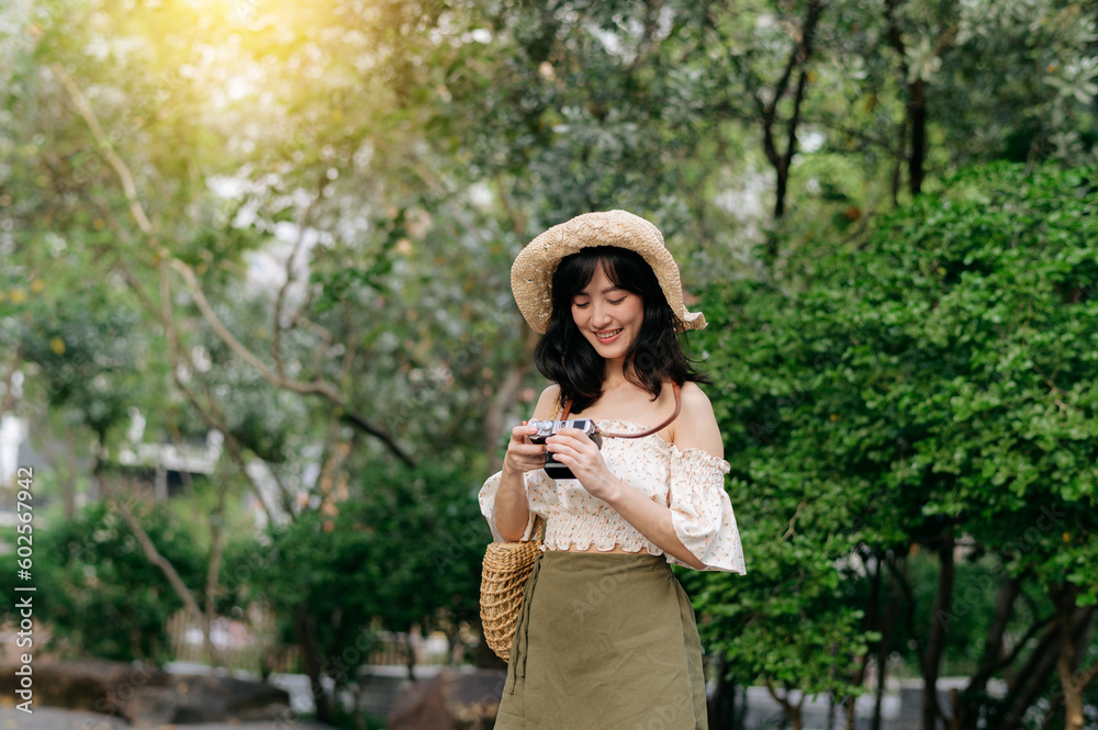 Portrait of asian young woman traveler with weaving hat and basket and a camera on green public park nature background. Journey trip lifestyle, world travel explorer or Asia summer tourism concept.