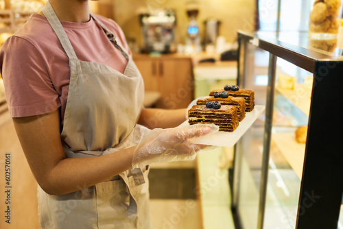 Cropped shot of young clerk in apron holding board with three fresh cakes or desserts while putting them or taking out of display in cafeteria