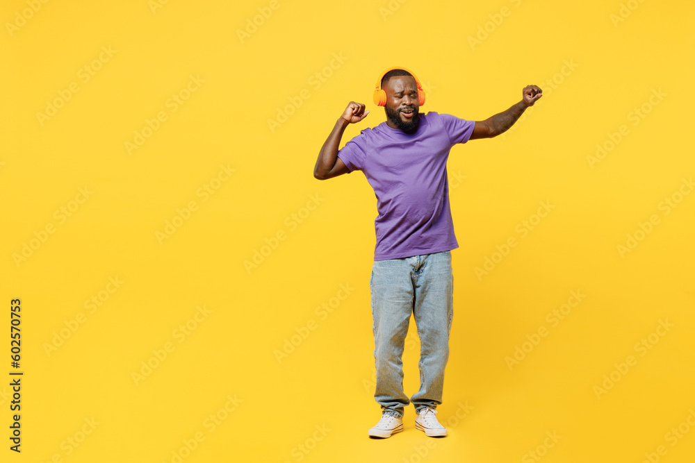Full body young man of African American ethnicity he wears casual clothes purple t-shirt headphones listen to music raise up hands dance isolated on plain yellow background studio. Lifestyle concept.
