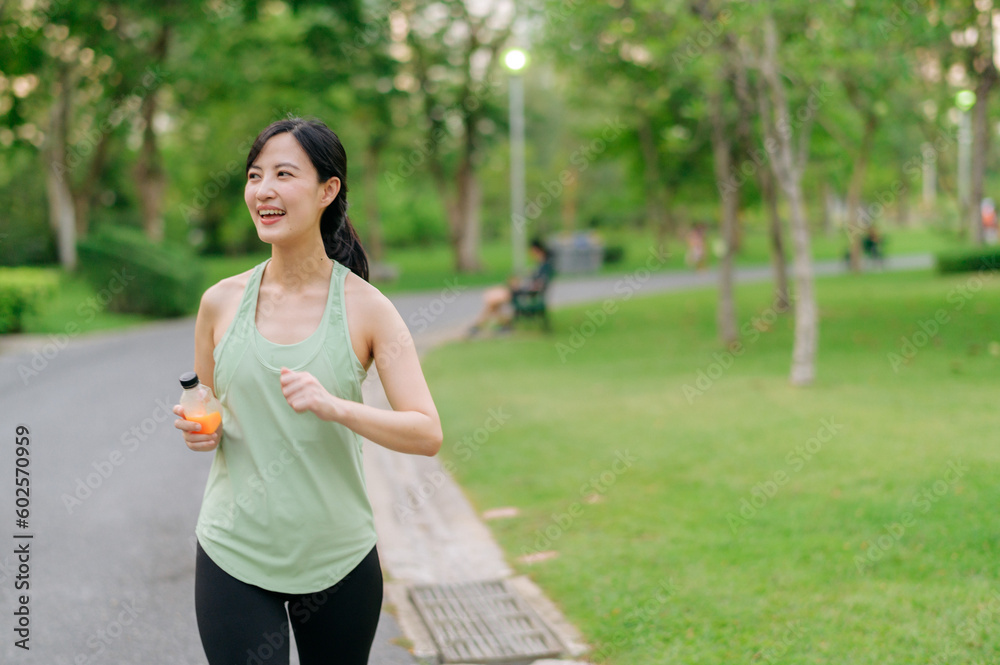 Female jogger. Fit young Asian woman with green sportswear drinking organic orange juice after running and enjoying a healthy outdoor. Fitness runner girl in public park. Wellness being concept Female jogger. Fit young Asian woman with green sportswear drinking organic orange juice after running and enjoying a healthy outdoor. Fitness runner girl in public park. Wellness being concept