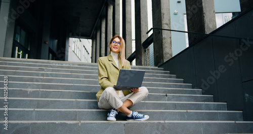Portrait of cheerful young businesswoman working online with laptop sitting on stairs outside the business center. Business or education concept.