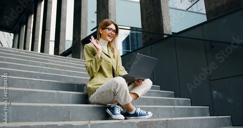 Smiling woman with laptop having online video chat while sitting on the stairs. Girl freelancer talking on video conference online outdoors.