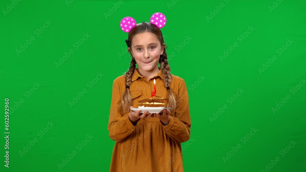 Positive beautiful teen girl with piece of birthday cake making wish blowing out candle looking at camera. Smiling confident Caucasian teenager celebrating holiday posing on green screen background