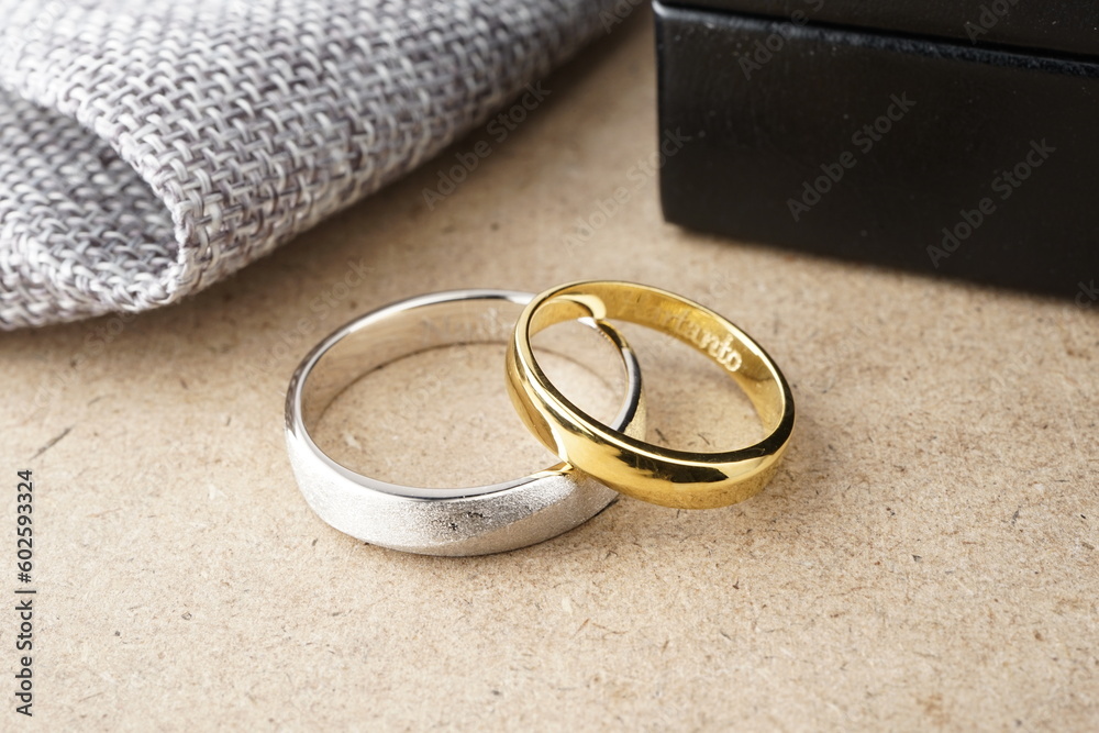 Couple gold and palladium wedding rings on a wooden pattern plinth and black ringbox behind with burlap trim Couple gold and palladium wedding rings on a wooden pattern plinth and black ringbox behind with burlap trim