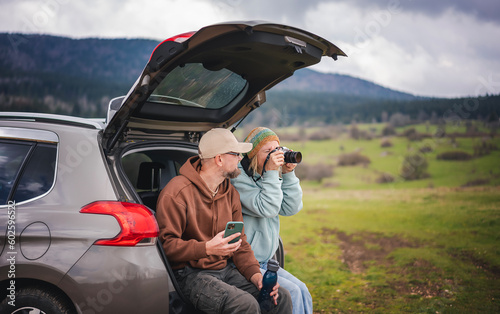 Romantic middle age caucasian couple traveling by car sitting in open trunk with photo camera and smartphone enjoying nature