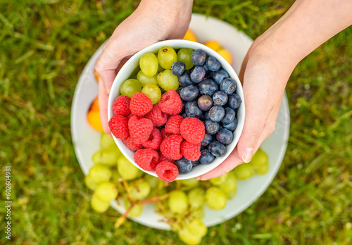 Miseczka z borówkami winogronem i malinami trzymana w rekach na tle talerza z owocami na zielonym tłe