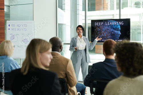 Confident young businesswoman in elegant suit making presentation of deep learning course while standing in front of audience at seminar