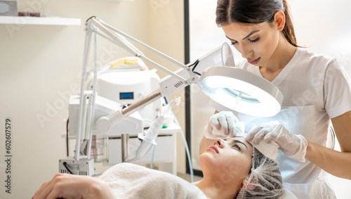 Slika na platnu Beautician squeezing acne and pimples from young woman's nose using lamp and magnifying glass in beauty spa clinic