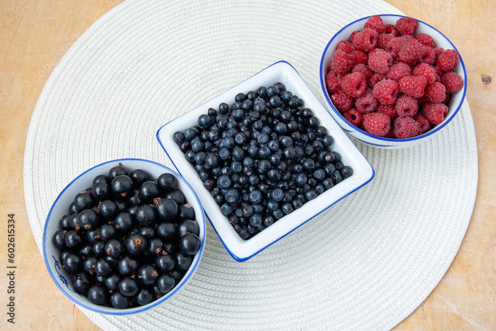 Three bowls with seasonal berries, a mix of fresh berries on a on a white round napkin on a wooden table, blueberries, currants, blueberries, raspberries Three bowls with seasonal berries, a mix of fresh berries on a on a white round napkin on a wooden table, blueberries, currants, blueberries, raspberries