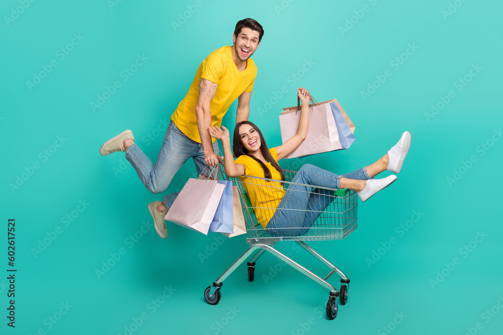 Photo of cool cheerful husband wife dressed yellow t-shirts driving cart rising shoppers isolated turquoise color background Photo of cool cheerful husband wife dressed yellow t-shirts driving cart rising shoppers isolated turquoise color background