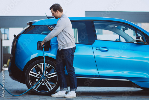 Young man charging electric car at home and using mobile phone