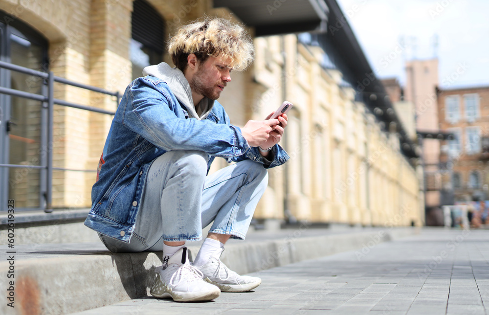 A young guy sits on the street and surfs the Internet using a smartphone. A young guy sits on the street and surfs the Internet using a smartphone.