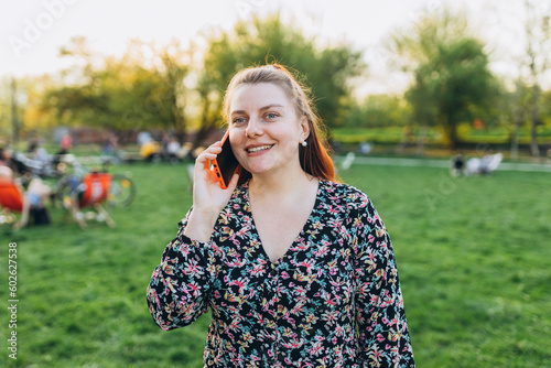 A portrait of a smiling beautiful woman talking on phone on nature background. Happy woman in dress is using a smartphone in park outdoors, summer time. Traveler