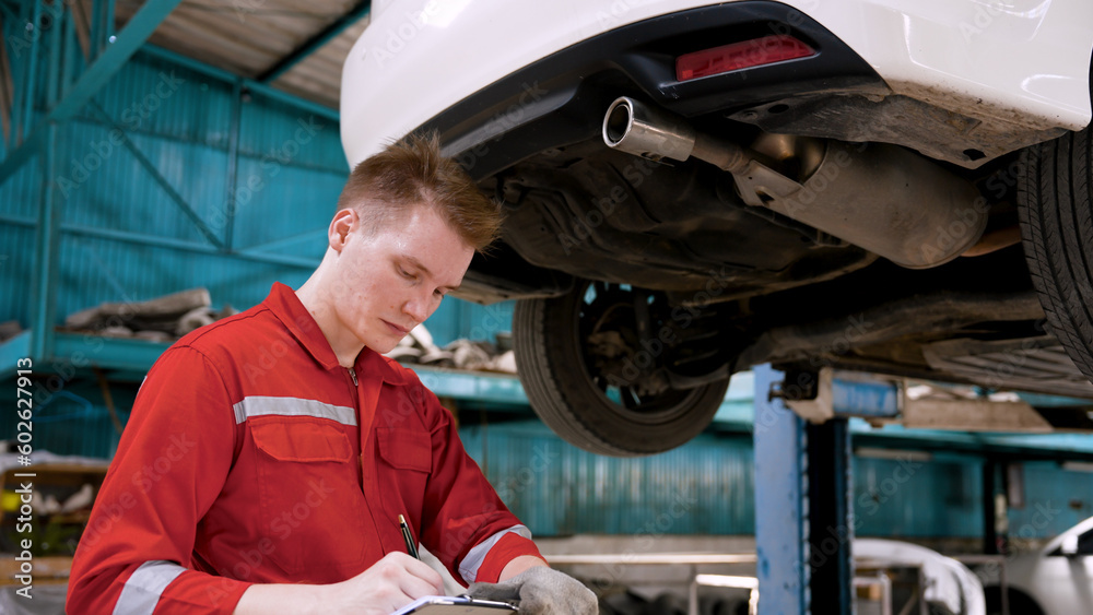 Man car mechanic Caucasian man, Checking front wheels raised car repair worn parts or part that has driving problems, In hand, held vehicle check book wrote note on problematic part. Man car mechanic Caucasian man, Checking front wheels raised car repair worn parts or part that has driving problems, In hand, held vehicle check book wrote note on problematic part.